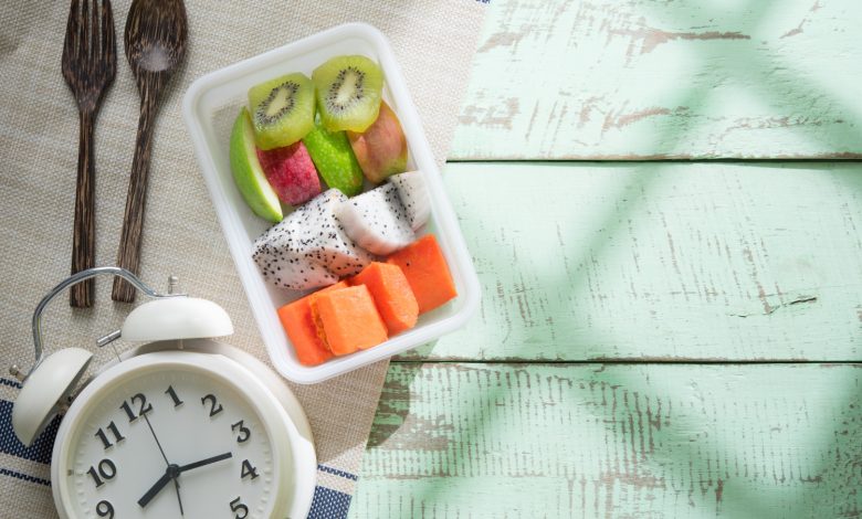 Tazón de frutas, cubiertos y reloj sobre mesa de madera, haciendo referencia a al horario de comidas